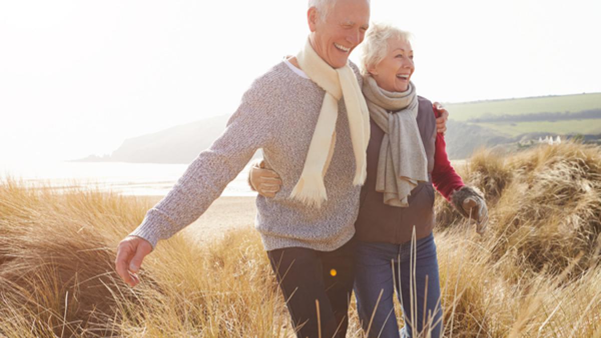 Senior Couple Walking Through Sand Dunes On Winter Beach Smiling